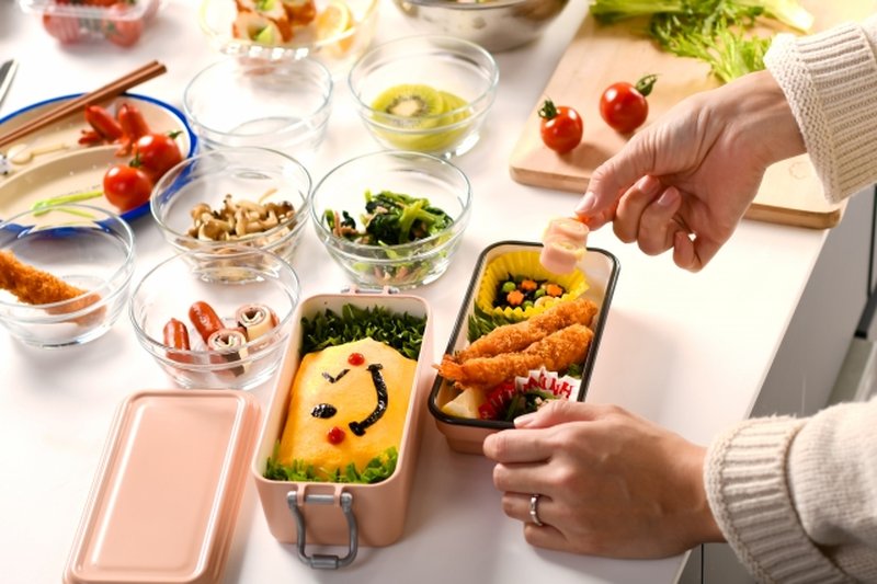 A parent assembling a large bento surrounded by many prepared ingredients — the kind of preparation typical the evening before undokai in Japan