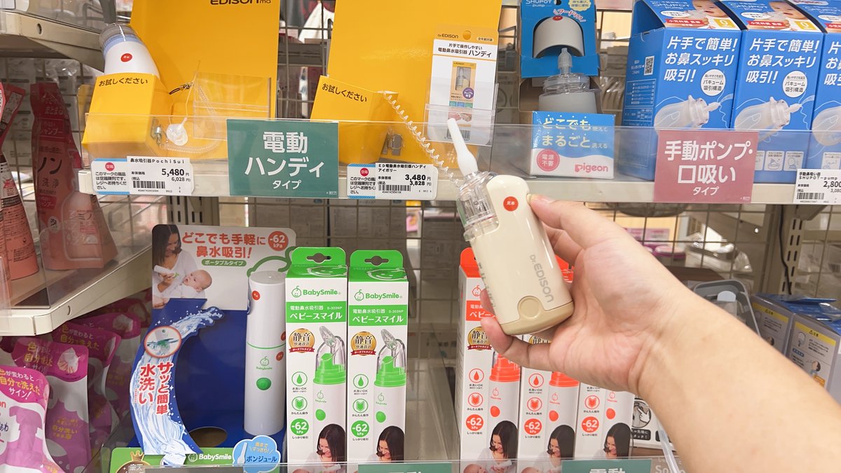 A hand holding a compact electric nasal aspirator in a Japanese baby shop, with shelf labels showing electric handy type and manual or mouth-suction type categories